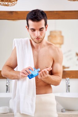 Omuzlarında havlu olan genç sporcu eline losyon döker, banyo aynasının önünde durur.