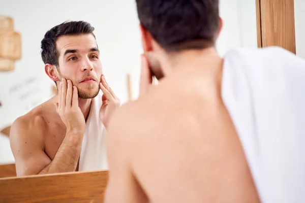 Brunet man lathers his face and stands with towel on his shoulders in front of bathroom mirror — Stock Photo, Image