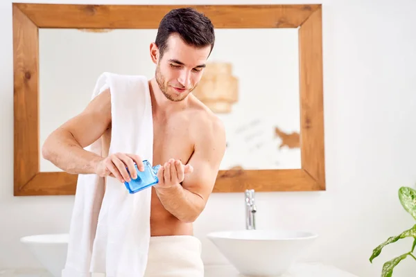 Masculino com toalha em seus ombros com loção na mão fica na frente do espelho do banheiro — Fotografia de Stock