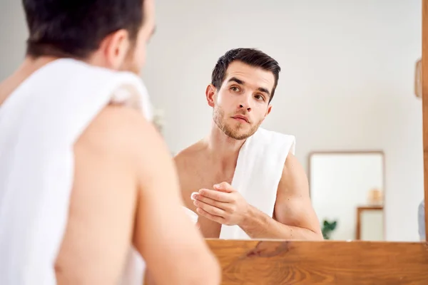 Brunet male spreads his face with white cream while standing in front of mirror in bath — Stock Photo, Image