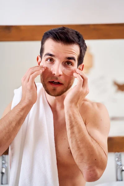 Brunet male applies transparent patches under his eyes while standing in bath opposite mirror — Stock Photo, Image