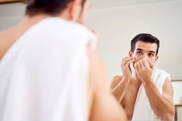 Young male applies transparent patches under his eyes while standing in bath opposite mirror — Stock Photo, Image