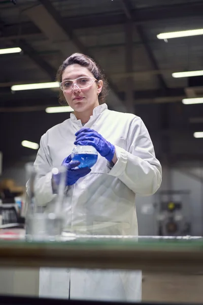 Brunetka laboratoryjna patrząc na kamerę w okularach i białym płaszczu z kolbą doświadczalną z niebieskim płynem w dłoniach prowadzi eksperymenty — Zdjęcie stockowe