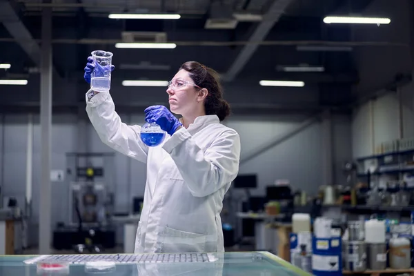 Chica de laboratorio en gafas y abrigo blanco con frasco con líquido azul en sus manos lleva a cabo experimentos —  Fotos de Stock