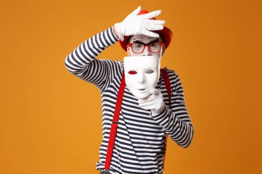 Kırmızı şapkalı ve yelekli pandomim palyaçosu, turuncu arka planda beyaz maske.