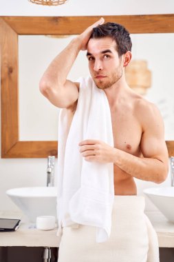 Brunet male with white towel on his shoulder standing by mirror in bath.