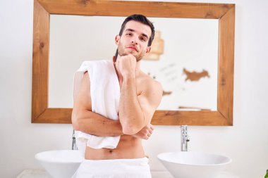 Pensive man with towel on shoulder stands near mirror in bath