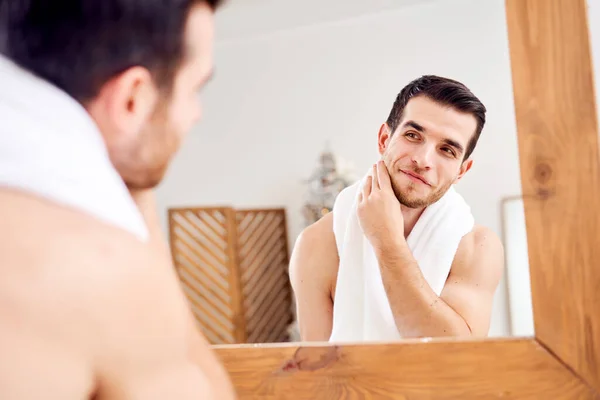 Unshaven undressed brunet with white towel on his neck standing near mirror — Stok fotoğraf