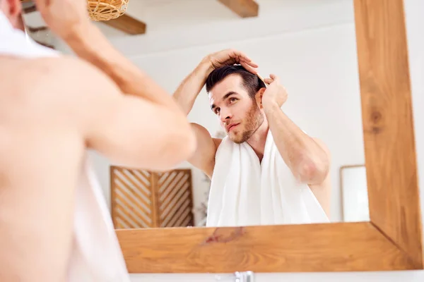 Молодой человек гладит волосы, стоя перед зеркалом — стоковое фото