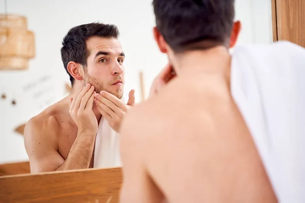 Brunet male lathers his face and stands with towel on his shoulders in front of bathroom mirror — Stock Photo, Image