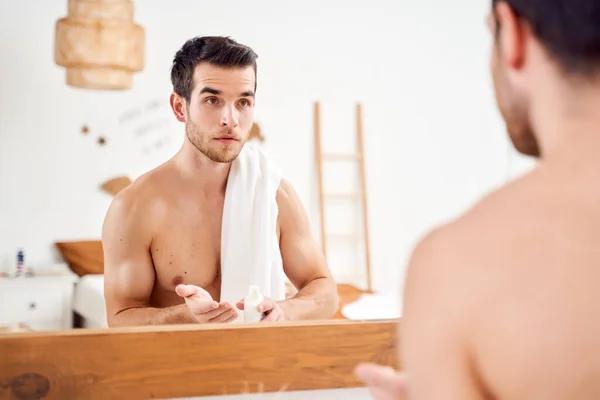 Young sports male with lotion in hands and towel on shoulders stands in front of bathroom mirror — Stockfoto