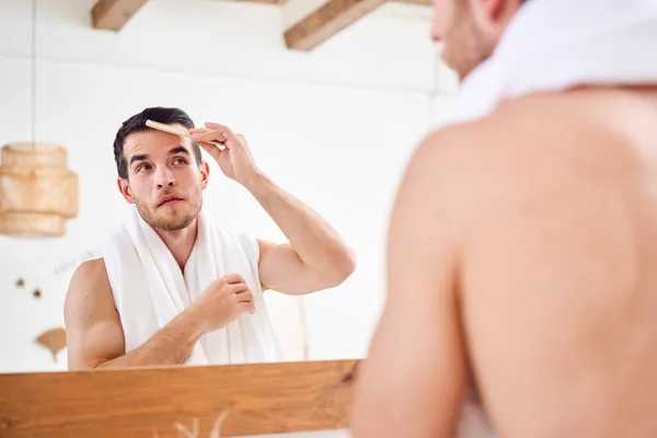 Joven hombre sin afeitar peinándose el pelo mientras está de pie con una toalla blanca en el cuello cerca del espejo en el baño —  Fotos de Stock