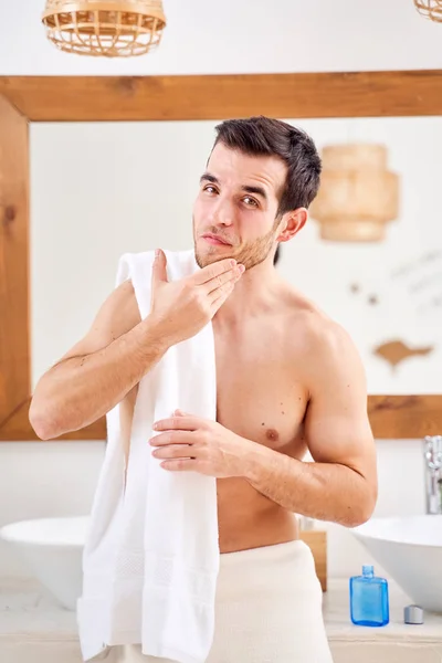 Male washes his face and stands with towel on his shoulders in front of bathroom mirror — Stock Photo, Image