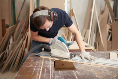 Carpenter in protective headphones and white gloves works on jigsaw in workshop