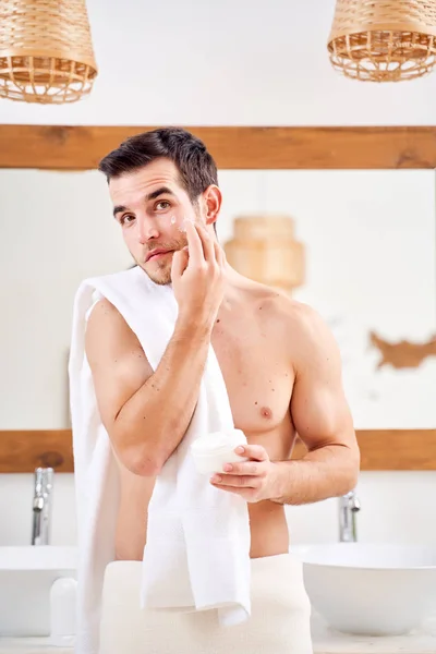 Young man spreads his face with white cream while standing in front of mirror in bath — Stock Photo, Image