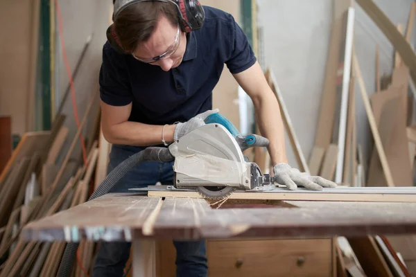 Young man in safety glasses and white gloves works on jigsaw in workshop — Stockfoto