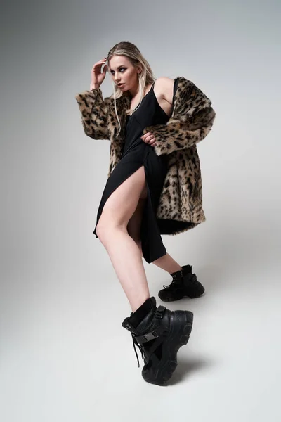 Blonde girl in leopard coat and boots looking at side while standing in studio — Stock Photo, Image