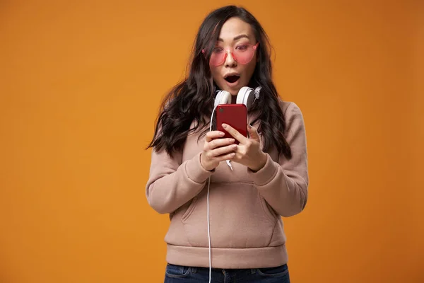 Aziatische brunette in roze bril met telefoon in haar handen geïsoleerd in studio — Stockfoto