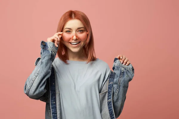 Mujer feliz en gafas rosas y chaqueta de mezclilla de pie en el estudio — Foto de Stock