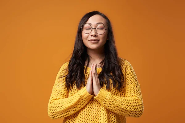Oración joven asiática mujer con los ojos cerrados con gafas y en suéter de punto amarillo sobre fondo naranja vacío — Foto de Stock