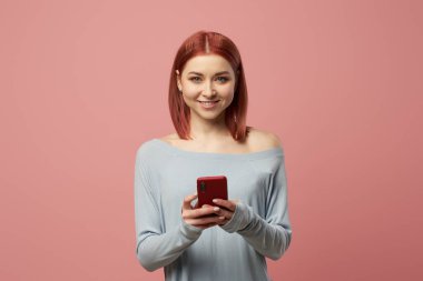 Elinde telefonuyla gülümseyen kızıl kadın izole edilmiş pembe arka planda stüdyoda duruyor.