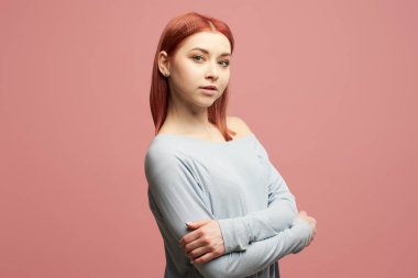 Stüdyoda kollarını kavuşturmuş, gri kazaklı, gülümseyen kızıl kadın.