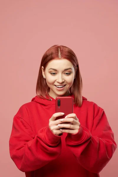 Молода імбирна жінка в червоному светрі з телефоном в руках ізольована на рожевому фоні — стокове фото