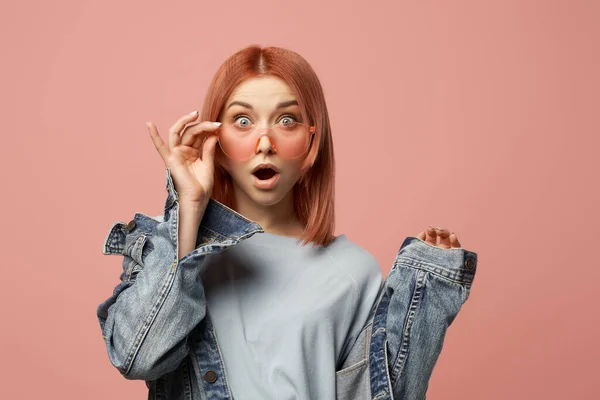 Överraskad kvinna i rosa glasögon och jeansjacka stående i studio — Stockfoto
