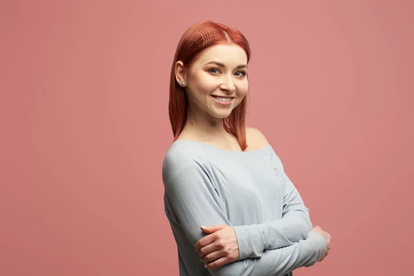 Smiling ginger girl in gray sweater with arms crossed standing in studio isolated on pink background — ストック写真