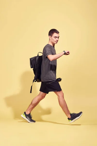Vista lateral del joven atleta con mochila mirando el reloj en su mano en el estudio — Foto de Stock
