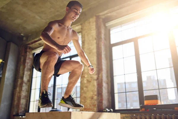 Zijaanzicht van springende atleet man op houten doos in de sportschool tegen de achtergrond van een groot raam — Stockfoto