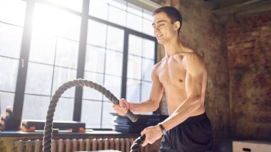 Spor salonundaki geniş pencerenin arka planında iplerle antrenman yapan genç bir atlet.