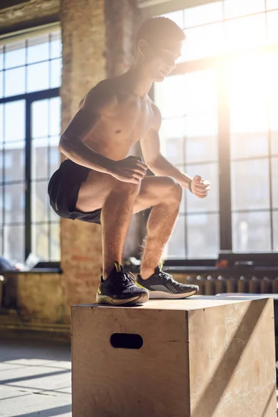 Side view of jumping young sportsman on wooden box in gym against background of large window — Stock Photo, Image