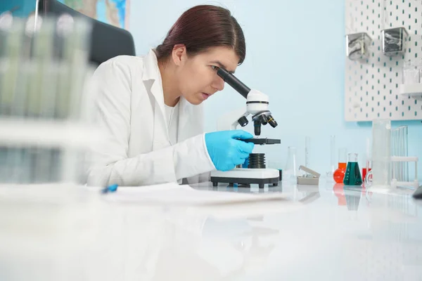 Brunette lab technicus kijkt naar microscoop in laboratorium. — Stockfoto