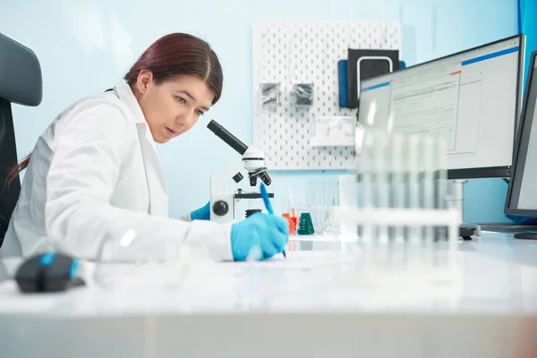 Jonge brunette lab technicus kijkt naar microscoop in laboratorium. — Stockfoto