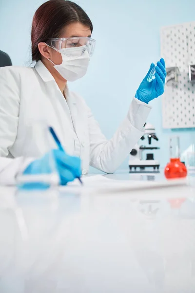 Young woman lab technician with test tube in hand makes note. — Stock Photo, Image