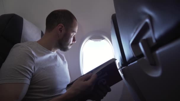 Mann sitzt am Fenster eines fliegenden Flugzeugs und liest Archivvideo des Magazins — Stockvideo