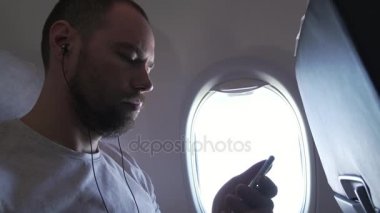 Uçak uçan ve müzik kulaklık smartphone ile pencere yanında oturan adam. stok görüntüleri video