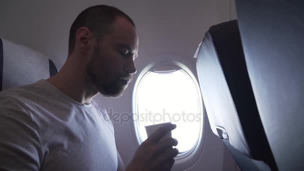 Mann sitzt am Fenster eines fliegenden Flugzeugs und trinkt einen Drink aus Pappbecher — Stockvideo