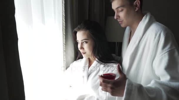 Aimant couple en manteaux blancs regarde le paysage à l'extérieur de la fenêtre, le gars apporte à la fille un verre de vin rouge vidéo de stock — Video