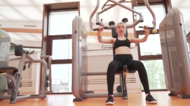 Νεαρό κορίτσι προπονητές τους μυς των χεριών σε ένα προσομοιωτή στο γυμναστήριο πλάνα βίντεο — Αρχείο Βίντεο