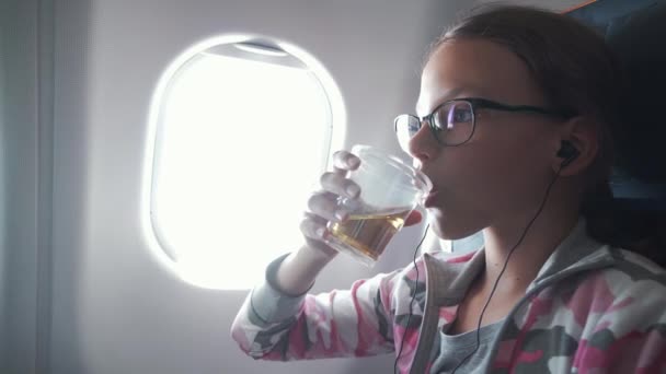 Ung flicka med glasögon och hörlurar klockor video på monitorn inbyggd i fåtölj och dricka juice i kabinen av flygplan arkivfilmer video — Stockvideo