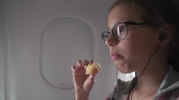 Девушка в очках и наушниках смотрит видео на мониторе, встроенном в кресло, и съедает рулон в кабине самолета — стоковое видео