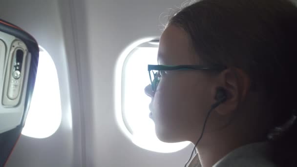 Junges Mädchen mit Brille und Kopfhörer sieht Video auf dem Monitor, der in Sessel in der Flugzeugkabine eingebaut ist — Stockvideo