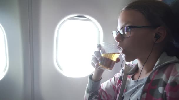 Ung flicka med glasögon och hörlurar klockor video på monitorn inbyggd i fåtölj och dricka juice i kabinen av flygplan arkivfilmer video — Stockvideo