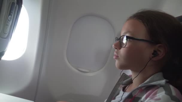 Junges Mädchen mit Brille und Kopfhörer sieht Video auf dem Monitor, der in Sessel in der Flugzeugkabine eingebaut ist — Stockvideo