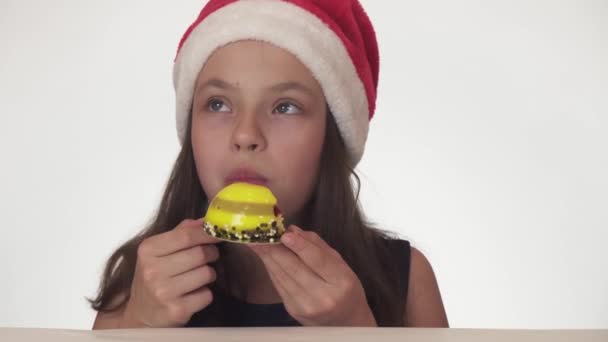 Hermosa chica adolescente en el sombrero de Santa Claus con un apetito y placer comiendo un pastel de cumpleaños en el fondo blanco material de archivo de vídeo — Vídeos de Stock