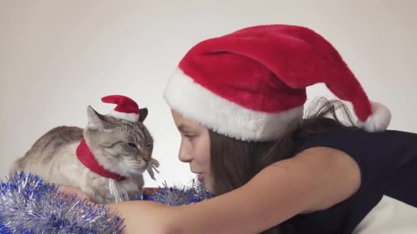 Beautiful teen girl and her cat in Santa Claus caps joyfully kissing on white background stock footage video — Stock Video