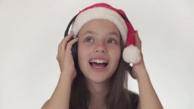 Güzel yaramaz kız genç bir Noel Baba şapkalı kulaklıklar müzik dinler ve boyunca üzerinde beyaz arka plan stok görüntüleri video şarkı söylüyor.