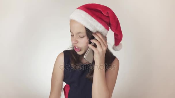 Beautiful naughty girl teenager in a Santa Claus hat emotionally communicates on the smartphone on white background stock footage video. — Stock Video